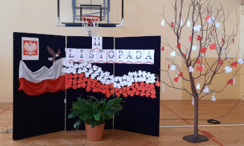 Sala gimnastyczna. Na ciemnej ściance napis 11 listopada. Godło i flaga Polski. Obok drzewko z białymi i czerwonymi liśćmi.