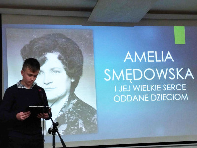 Uczeń czyta przez mikrofon. W tle wizerunek i napis Amelia Smędowska i jej wielkie serce oddane dzieciom