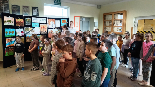 Uczestnicy szkolnego konkursu iteracko-plastycznego stoją na tle nagrodzonych i wyróżnionych konkursowych prac plastycznych.