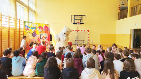 Dzieci siedzą w sali gimnastycznej, oglądając występ osoby w stroju niedźwiedzia polarnego, na tle kolorowego baneru „Cyrk Szok”.