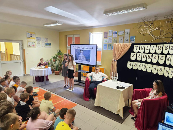 Uczniowie podczas akademii szkolnej upamiętniającej Juliusza Słowackiego, prezentujący inscenizację i recytujący wybrane utwory poety, otoczeni grupą słuchających dzieci.