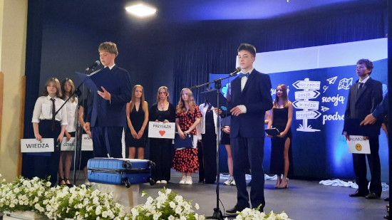 Uczniowie ubrani formalnie wygłaszają przemówienia podczas szkolnej uroczystości pożegnania absolwentów, trzymając kartki z hasłami takimi jak „Wiedza”, „Przyjaźń” i „Humor”.