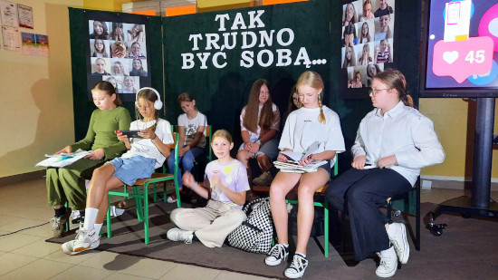 Grupa uczniów siedzi na scenie podczas przedstawienia „Tak trudno być sobą” Szkolnego Koła Teatralnego, z widocznym napisem tytułu za nimi.