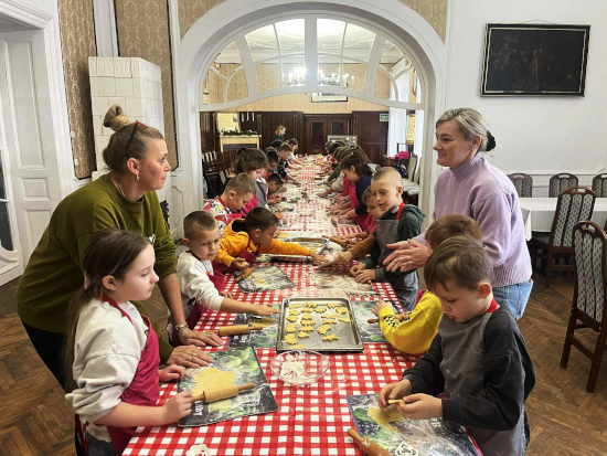 Dzieci siedzące przy długim stole z&nbsp;czerwoną, kratowaną ceratą formują i&nbsp;wykrawają świąteczne ciasteczka podczas warsztatów w&nbsp;dworku, nad którymi czuwają dwie dorosłe osoby.